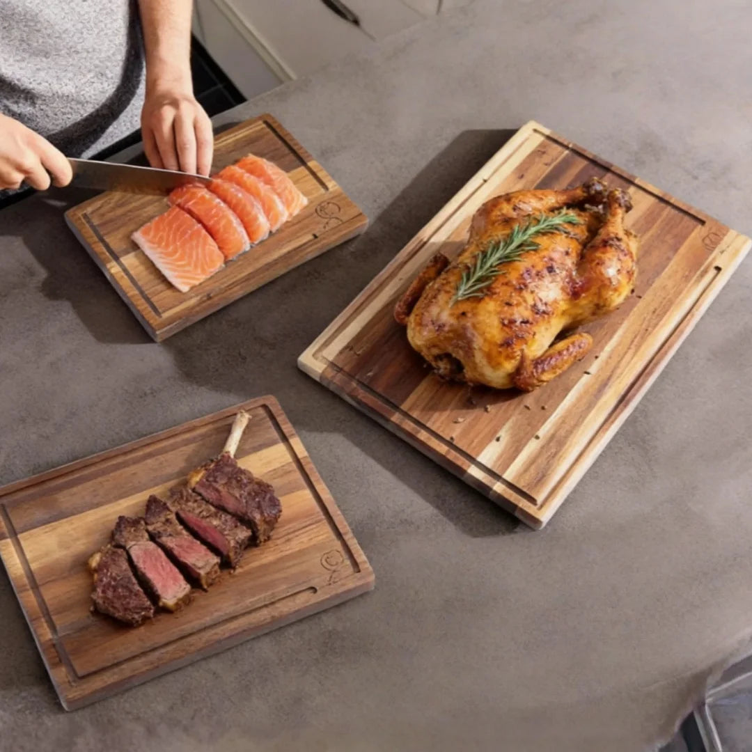 Set de 3 planches à découper en chêne avec rigole à jus Nesty, trois planches avec saumon tranché, poulet rôti au romarin et steak tranché, sur plan de travail gris