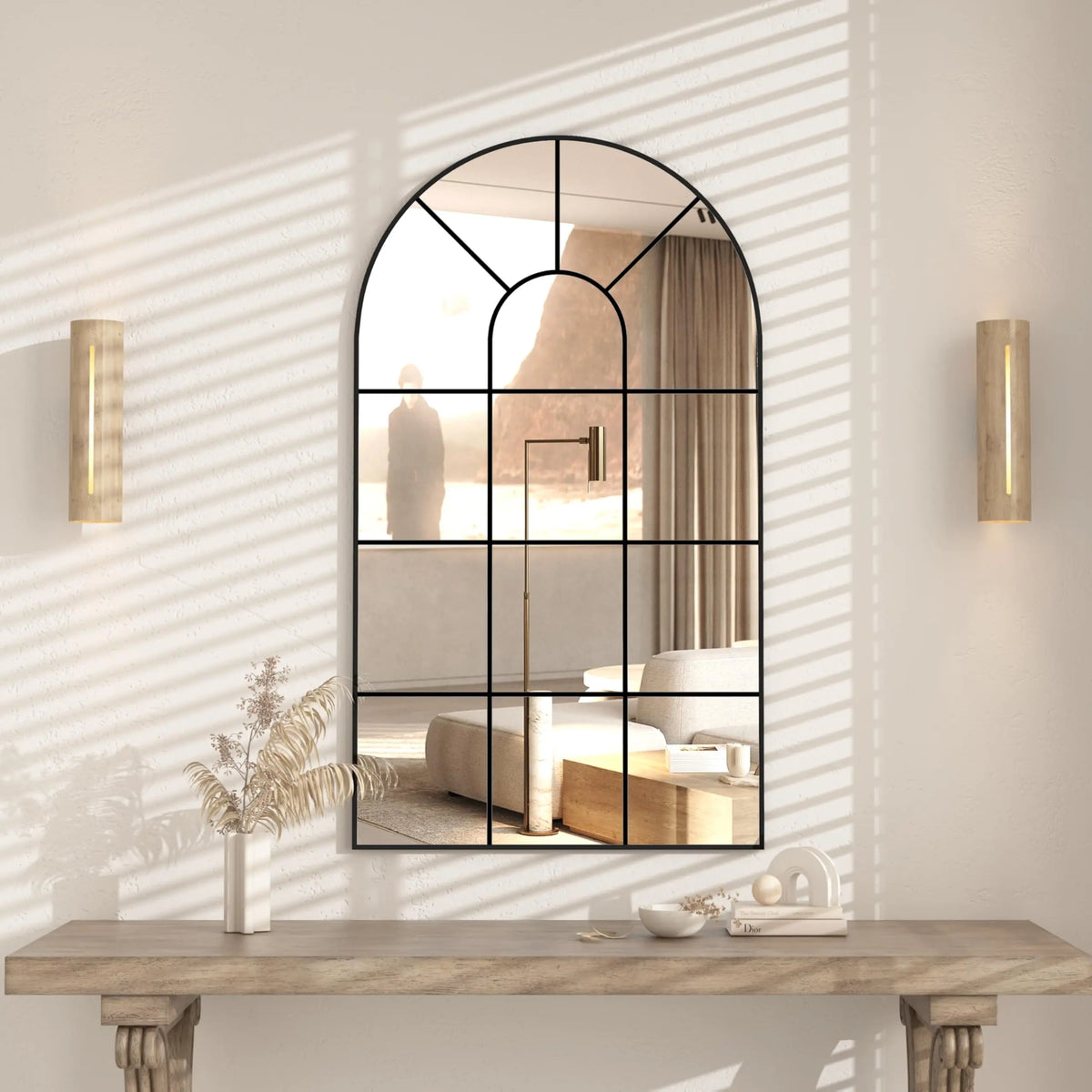 Miroir fenêtre cintré Nesty - Cadre noir rustique, accroché sur un mur beige baigné de lumière, au-dessus d'une console avec plante