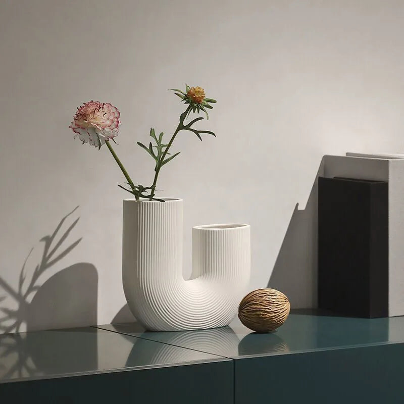 Vase céramique forme U, style nordique épuré - Nesty - Vase blanc en forme de U avec deux fleurs roses et jaunes, posé sur un meuble bleu près d'une boule décorative