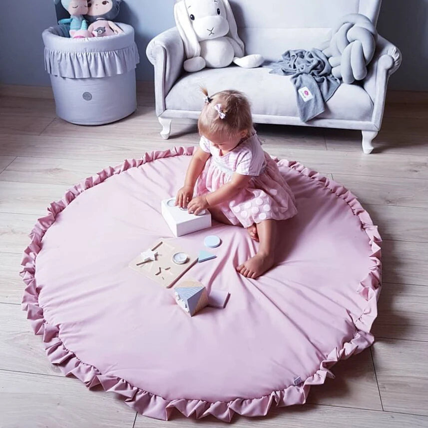 Tapis rond en coton pour bébé — coin jeux - Nesty, variante rose à bordure volantée, enfant assise en train de jouer avec des jouets en bois