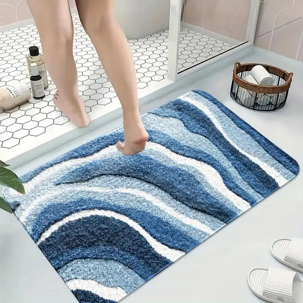 Tapis de bain à motif de vagues bleues, posé au sol près d'une douche à carrelage hexagonal, avec un pied posé dessus.