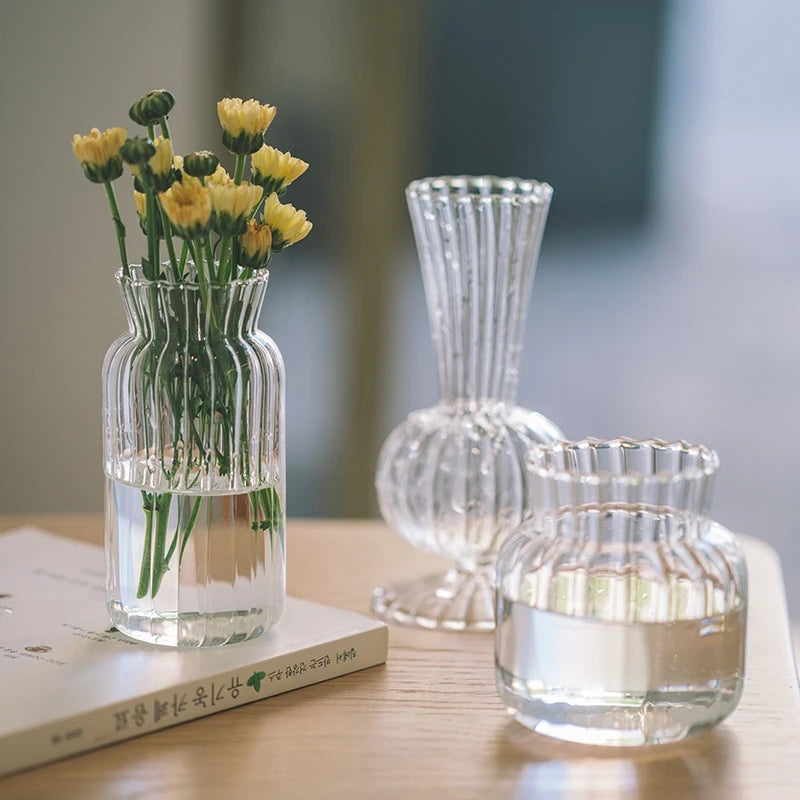 Vase en verre transparent strié, mini format rétro Nesty, trois formes côtelées transparentes, l'une garnie de fleurs jaunes posée sur un livre