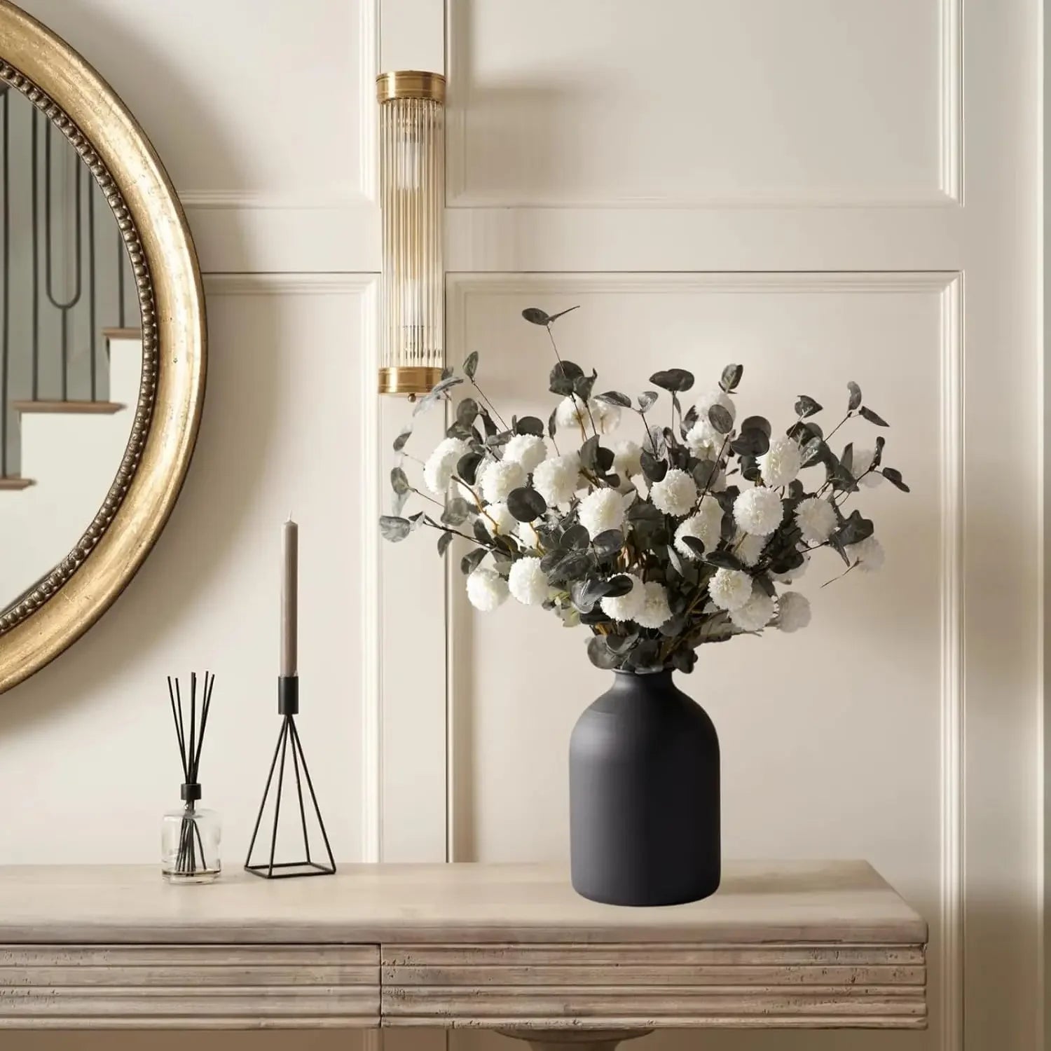 Vase en métal noir mat, finition sablée Nesty, cylindrique garni du même bouquet de boules blanches et eucalyptus, sur console beige avec miroir doré