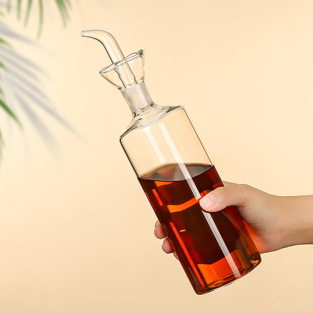 Burette à huile d'olive en verre borosilicate, anti-goutte Nesty, main tenant une burette en verre inclinée remplie d'huile ambrée, bec verseur relevé, fond beige avec feuillage
