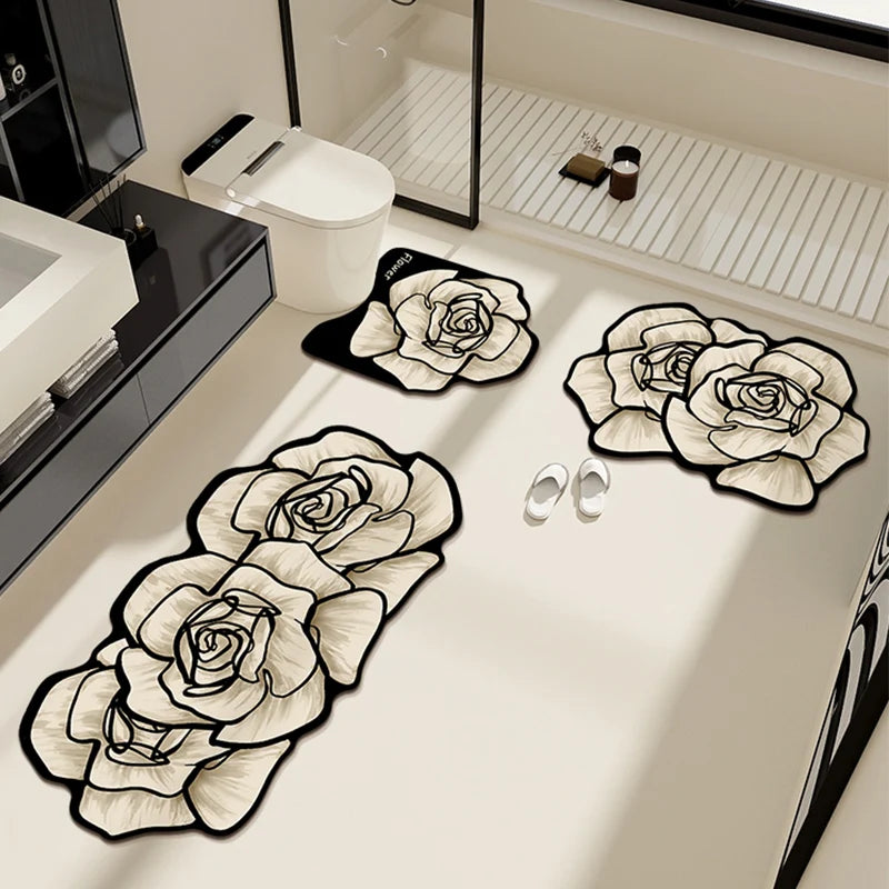 Set de trois tapis de salle de bain en forme de roses blanches à contour noir, disposés au sol autour d'une paire de chaussons, avec toilettes et douche en arrière-plan.