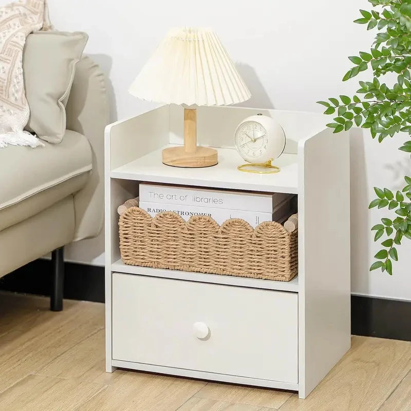 Panier de rangement en corde de papier tressée, bord festonné et poignées en bois Nesty, panier posé sur l'étagère d'une table de chevet blanche avec magazines, lampe et réveil