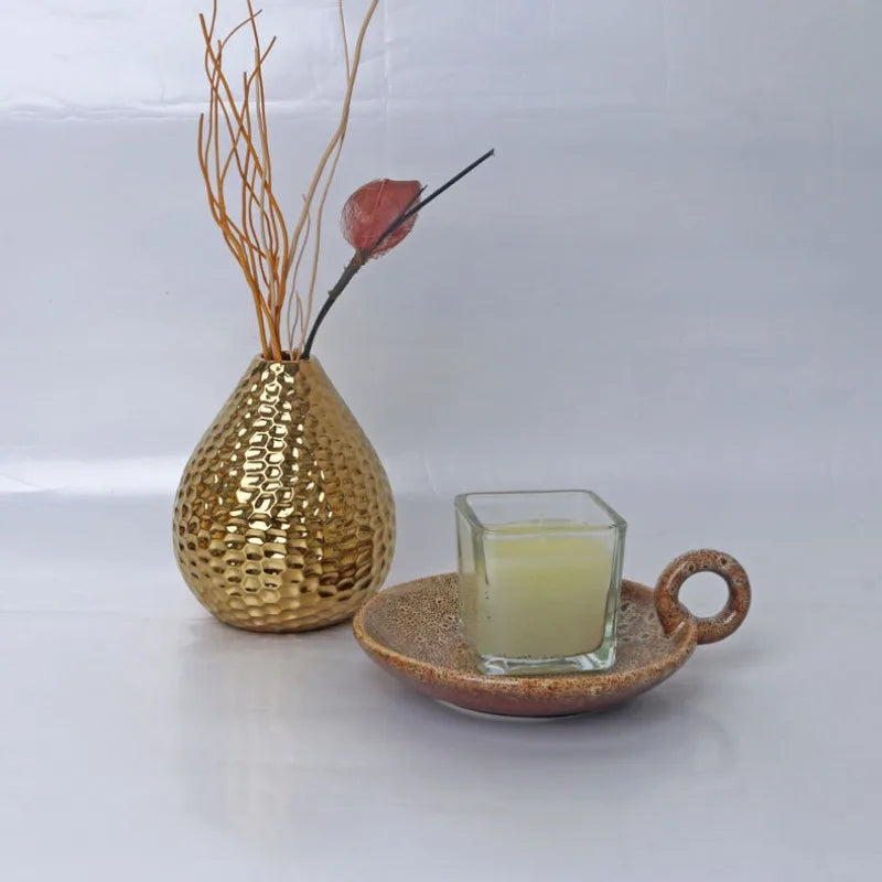 Bougeoir en céramique vieillie Nesty - Vase doré martelé avec branches séchées à côté d'une bougie en pot de verre posée sur la coupelle glaçurée