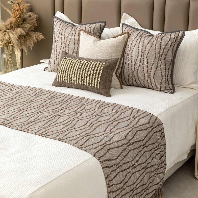 Housse de coussin en coton et lin beige, bordure texturée Nesty, deux housses de coussin à motif de lignes ondulées brunes et un coussin chiné beige à bordure marron, avec chemin de lit assorti, sur lit blanc