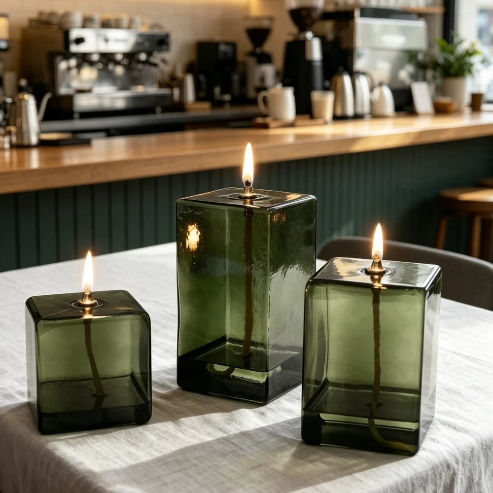 Lampe à huile en verre vert Nesty - Trois lampes cubiques allumées sur une table dans un café, comptoir avec machine à café en arrière-plan