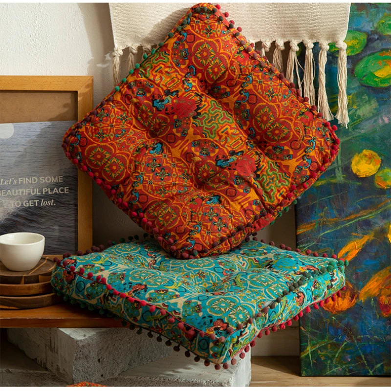 Gros coussin de sol en coton et lin épais, esprit marocain Nesty, coussin de sol carré orange à pompons et motif marocain, posé en losange sur un coussin turquoise assorti, devant un tableau