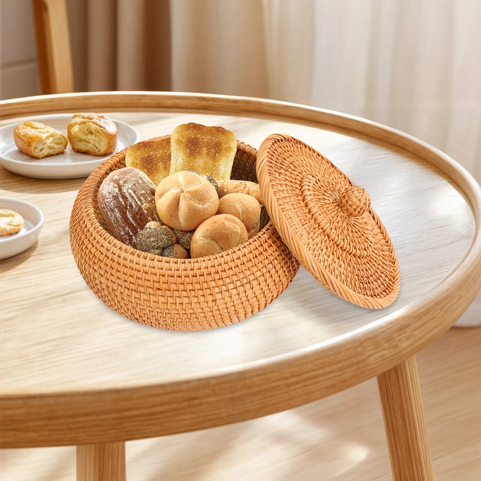 Panier de rangement artisanal en rotin naturel avec couvercle Nesty, panier ouvert garni de pains et petits pains, sur table ronde en bois, assiette de viennoiseries à côté