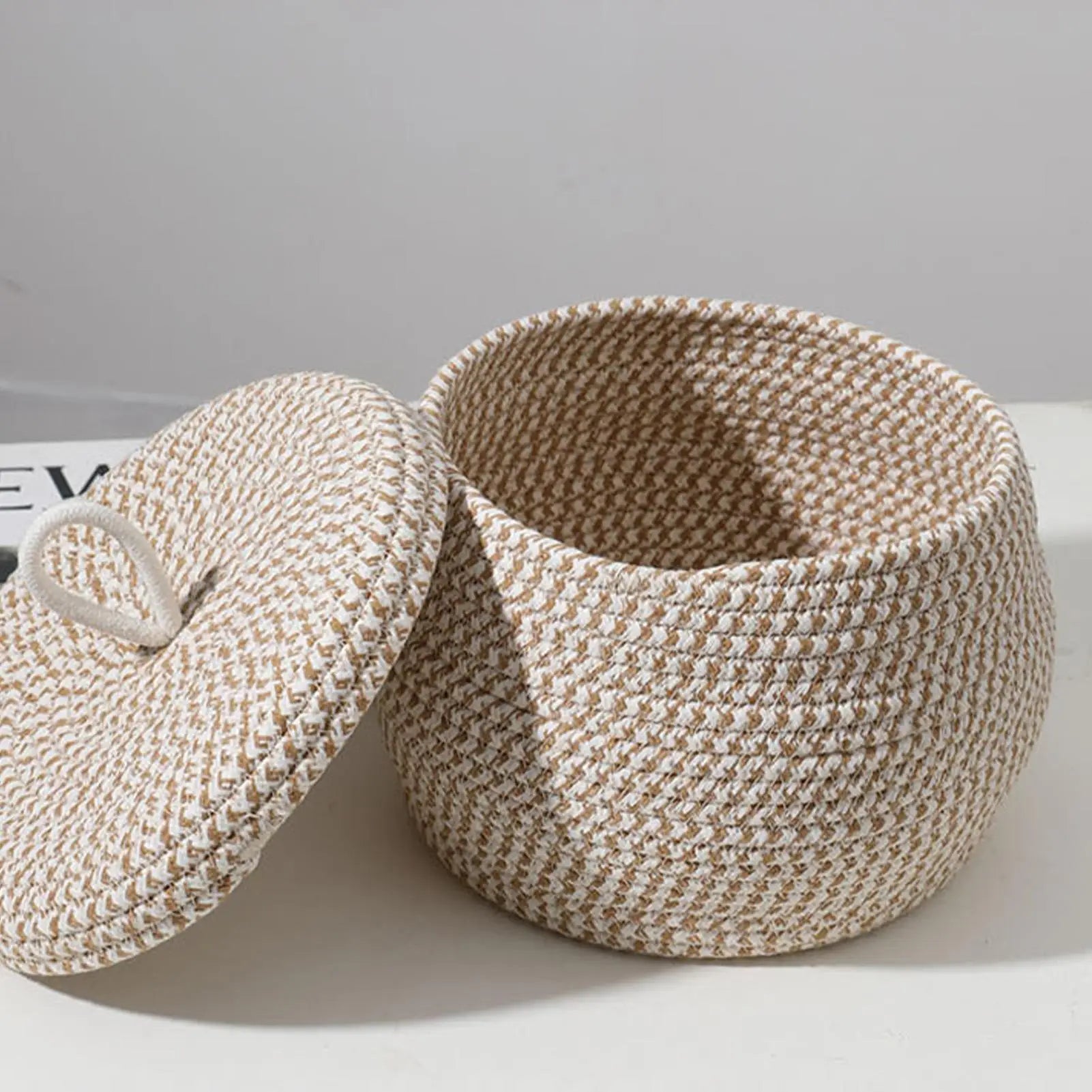 Petit panier rond tressé en corde de coton avec couvercle Nesty, panier vide avec couvercle posé contre lui, sur surface blanche avec livre