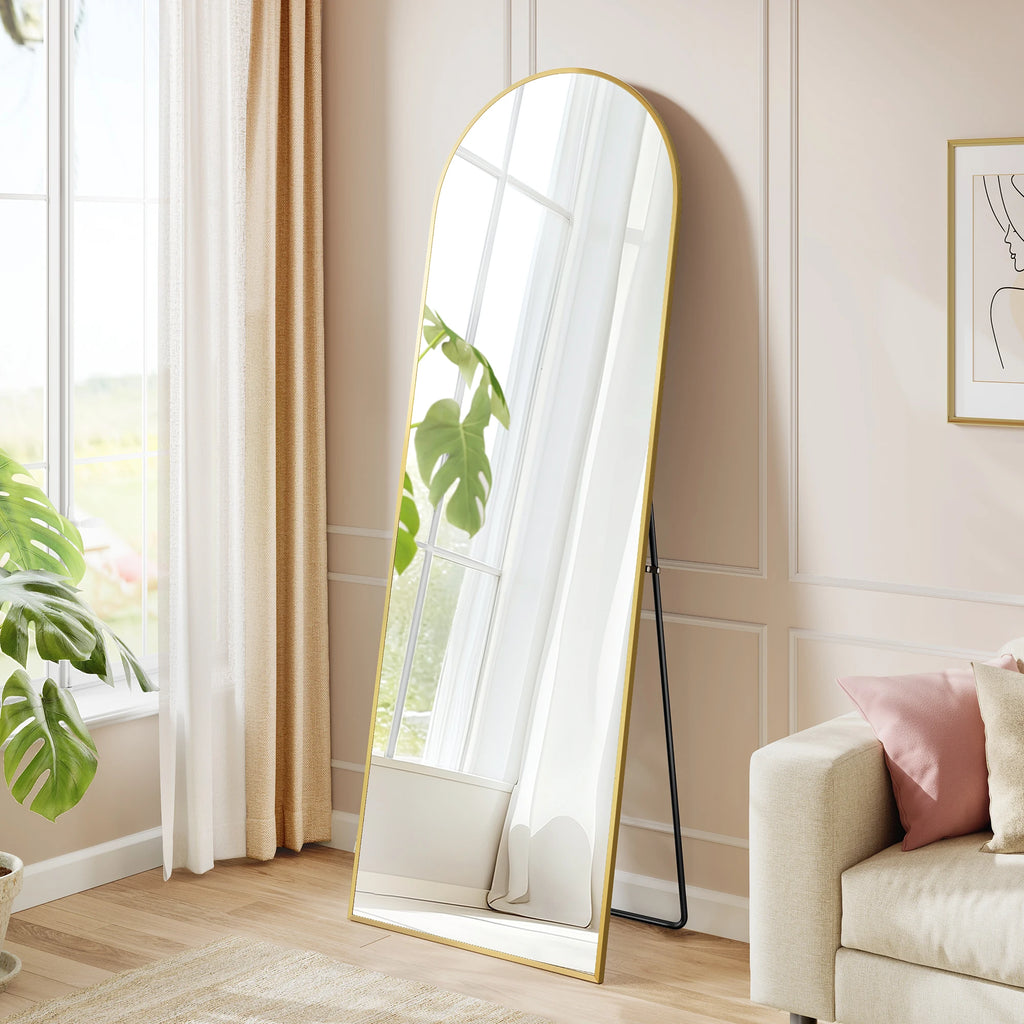 Miroir psyché cintré Nesty - Cadre doré posé au sol, reflet d'un rideau, d'une plante et d'un canapé à coussin rose