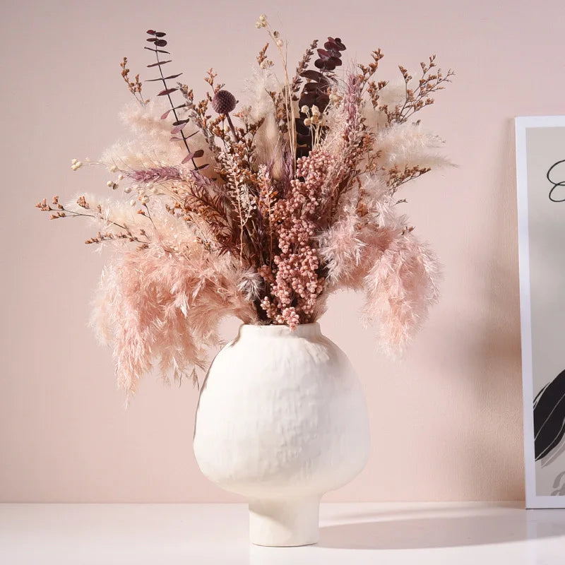 Vase en poterie fait main, grain brut, esprit artisanal Nesty, vase blanc texturé en forme de galet sur pied, garni d'un bouquet de pampas et fleurs séchées roses, sur fond rose poudré