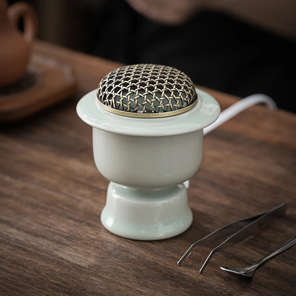 Brûle-parfum électrique en céramique Nesty - Modèle vert celadon avec couvercle grillagé doré, posé sur table en bois avec pinces