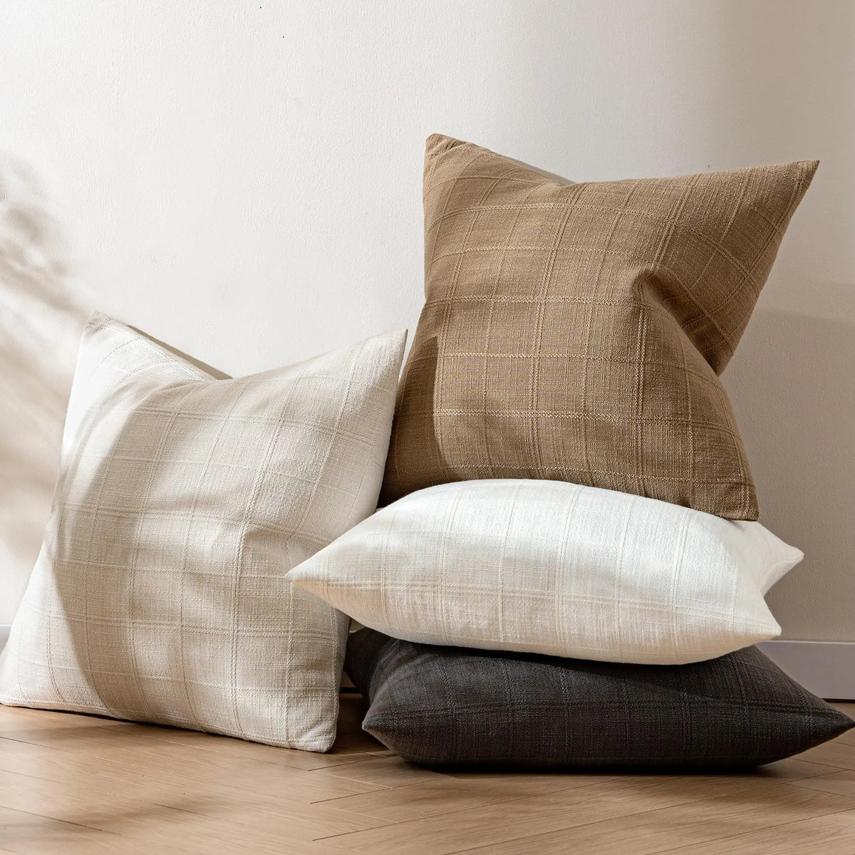 Housse de coussin en lin à carreaux, esprit campagne Nesty, quatre mêmes housses en lin à carreaux, coloris camel, écru, blanc cassé et anthracite, empilées au sol contre un mur