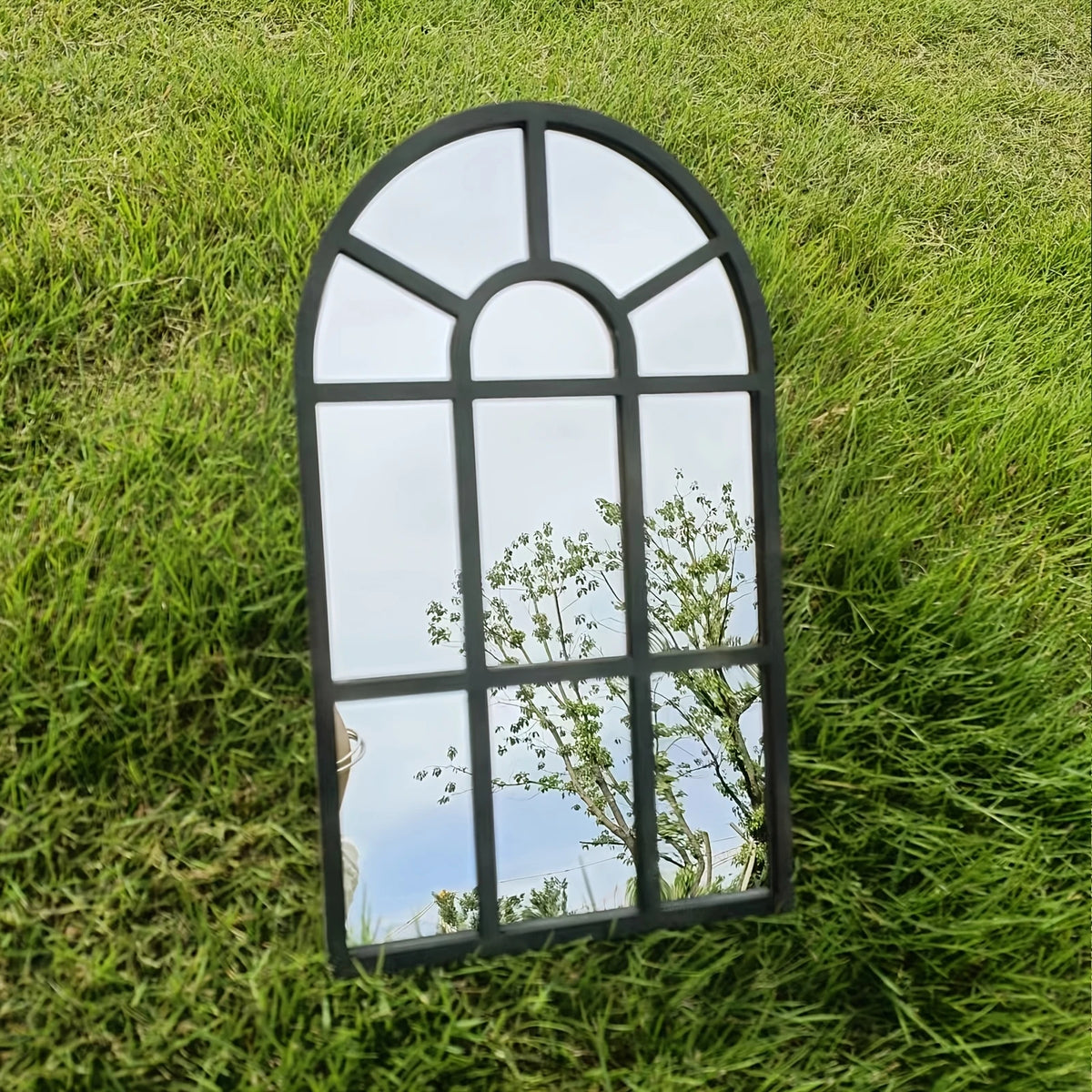 Miroir fenêtre en bois et acrylique Nesty - Format simple, miroir fenêtre cintré noir posé debout sur une pelouse, reflet du ciel et d'un arbre