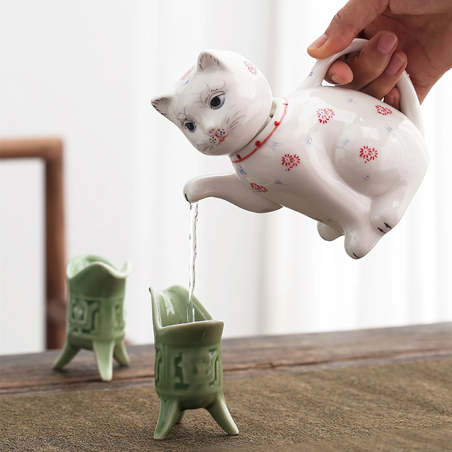 Théière en porcelaine forme de chat Nesty, en forme de chat blanc à motifs floraux, versant de l'eau depuis sa patte dans deux petits gobelets verts à pieds