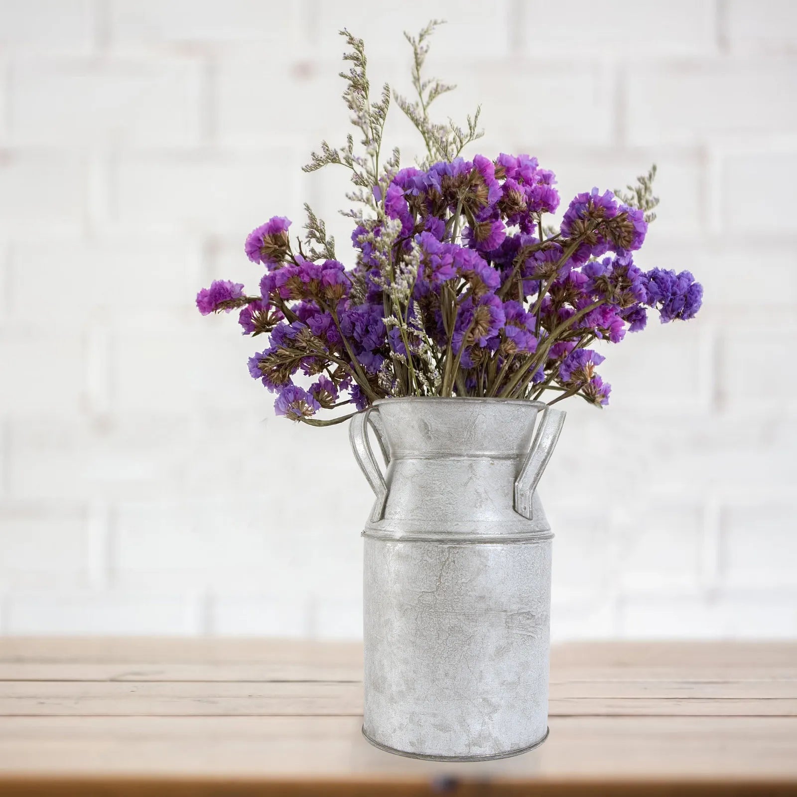 Vase seau en métal patiné Nesty - Avec fleurs de statice violettes, sur table en bois devant un mur en briques blanches