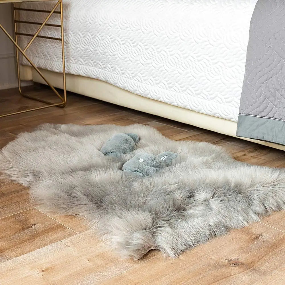 Tapis en fausse fourrure grise de forme irrégulière, posé au pied d'un lit sur un parquet, avec deux petites peluches en forme de chaussons dessus.