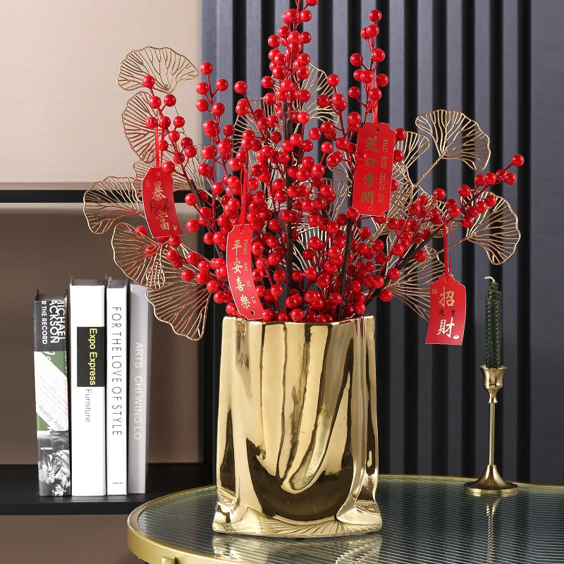 Vase en céramique argentée, forme sac plissé Nesty, version dorée effet froissé garnie de branches à baies rouges et feuilles dorées, sur table avec livres