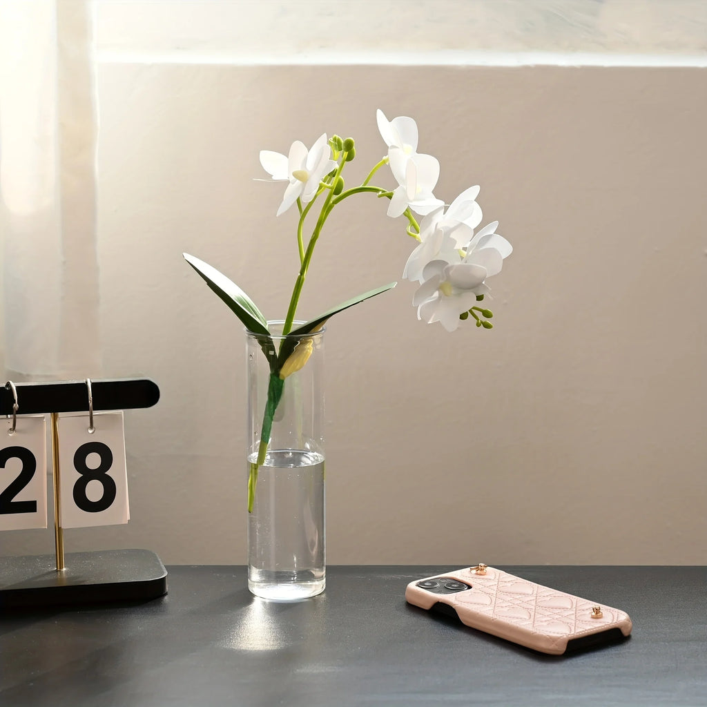Set de vases cylindriques transparents, trois tailles - Nesty - Vase garni d'une branche d'orchidée blanche, posé sur un bureau près d'un calendrier de bureau et d'une coque de téléphone rose