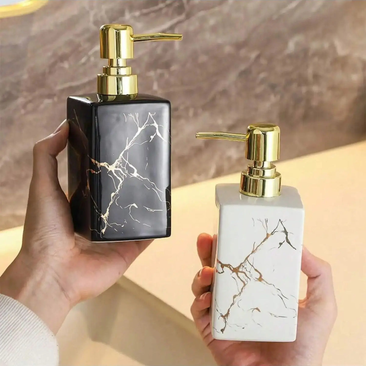 Distributeur de savon en céramique effet marbre Nesty, mains tenant les deux distributeurs de savon céramique marbré noir et blanc à pompe dorée