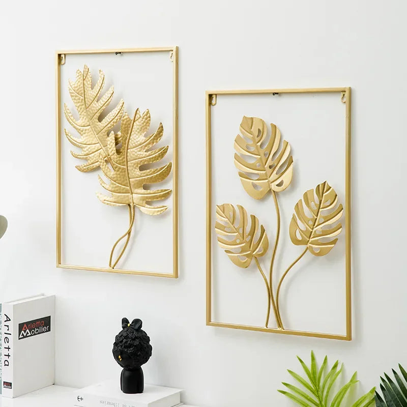 Pendentif mural en fer forgé à motif végétal Nesty, deux cadres dorés à motifs de fougère et de feuille de monstera sur un mur blanc, avec livres et figurine noire