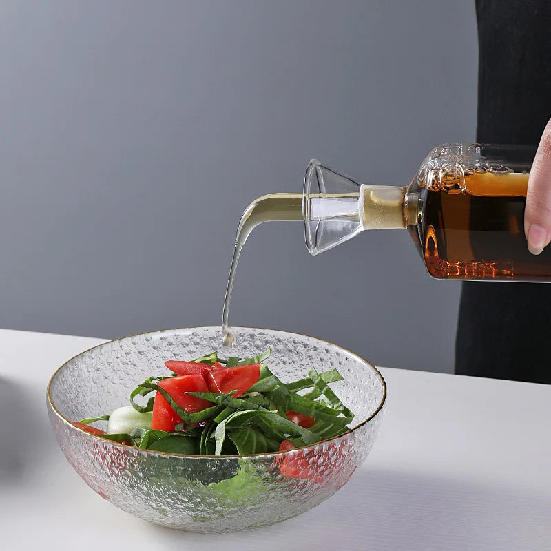 Pot à huile en verre borosilicate, anti-goutte Nesty, main versant un liquide ambré depuis la bouteille sur une salade dans un bol en verre texturé