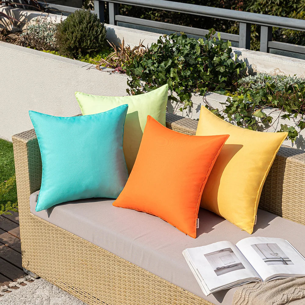 Housses de coussin extérieur imperméables, lot de 2 Nesty, cinq housses de couleurs variées turquoise, vert clair, orange et jaune posées sur un canapé en rotin extérieur