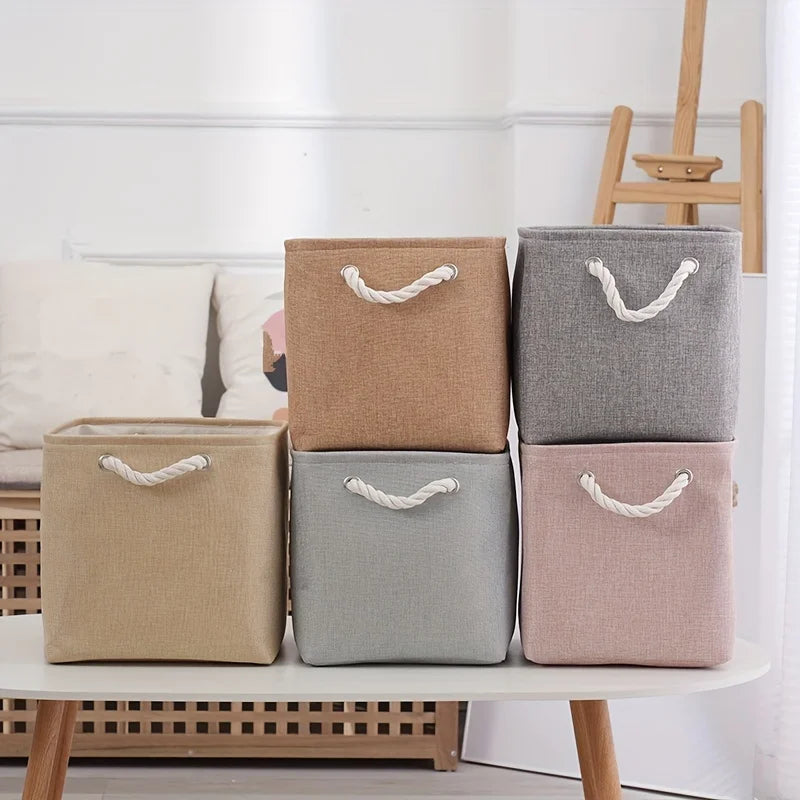 Boîte de rangement pliable en lin pour vêtements Nesty, six boîtes de couleurs et tailles différentes (beige, taupe, gris, vert d'eau, rose) sur une table ovale blanche, salon avec canapé