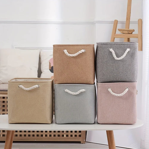 Boîte de rangement pliable en lin pour vêtements Nesty, six boîtes de couleurs et tailles différentes (beige, taupe, gris, vert d'eau, rose) sur une table ovale blanche, salon avec canapé