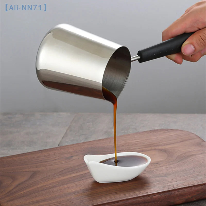 Casserole à manche long en inox pour lait et chocolat chaud Nesty, café versé depuis la casserole tenue en main dans une saucière blanche, sur planche en bois