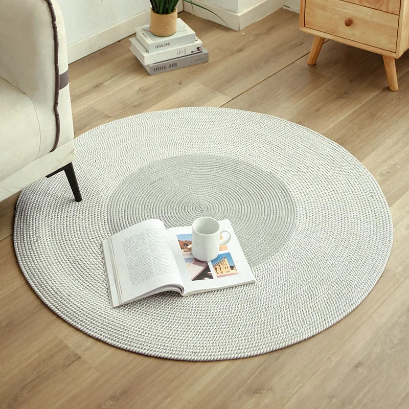 Petit tapis rond tissé à franges - Nesty, variante gris clair posée sur un parquet avec un livre et une tasse