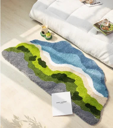 Tapis de chevet en forme libre représentant un paysage de rivière bleue serpentant entre des collines vertes et grises, posé au sol près d'un lit et d'un plateau petit-déjeuner.