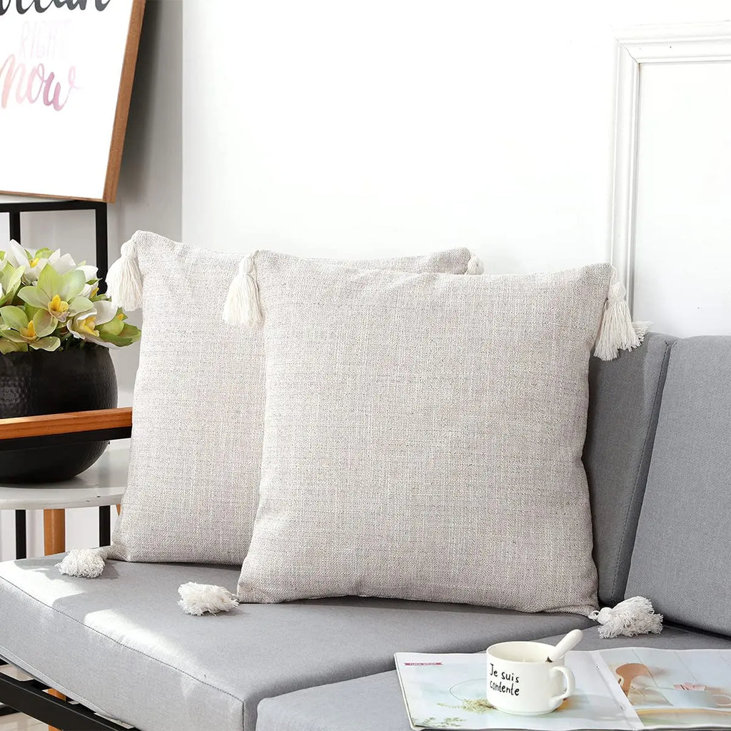 Housse de coussin en lin avec pompons, toutes saisons Nesty, deux housses de coussin beige clair à pompons blancs, posées sur canapé gris avec tasse et magazine