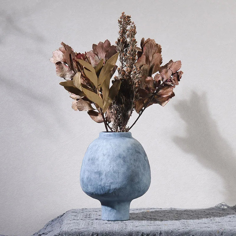 Vase en poterie fait main, grain brut, esprit artisanal Nesty, vase bleu grisé texturé en forme de galet sur pied, garni de feuillage séché brun et rose, sur nappe grise