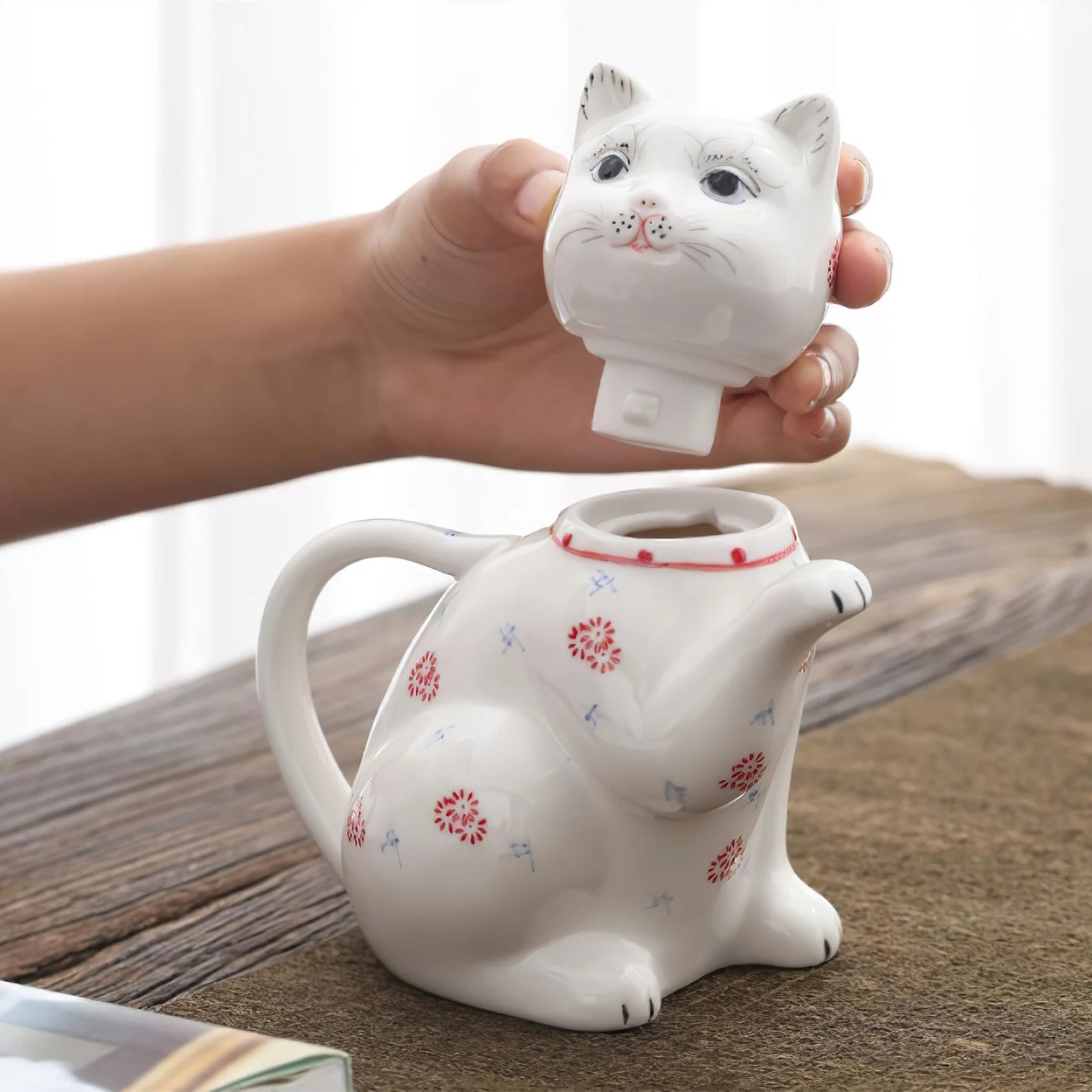 Théière en porcelaine forme de chat Nesty, tête du chat retirée en guise de couvercle, tenue en main au-dessus du corps ouvert