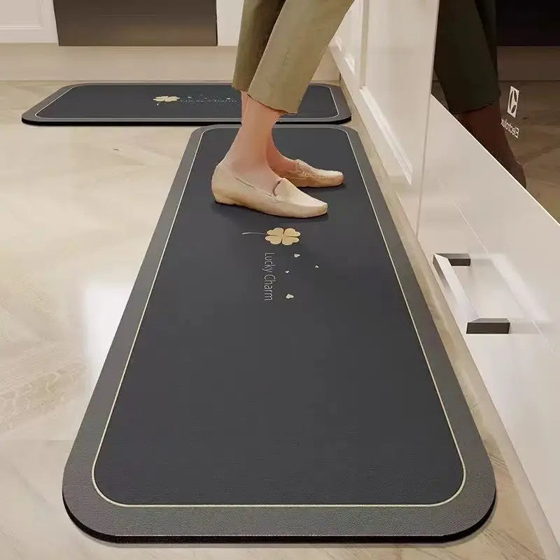 Tapis de cuisine long gris foncé à motif de trèfle doré 'Lucky Charm', posé au sol devant des meubles de cuisine blancs, avec une personne debout dessus.