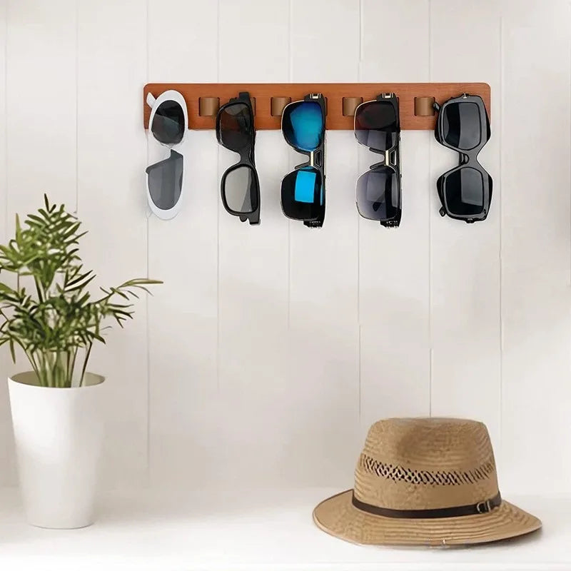 Porte-lunettes mural en bois Nesty, variante bois clair montée sur un mur lambrissé blanc, cinq paires de lunettes de soleil accrochées, plante et chapeau à proximité