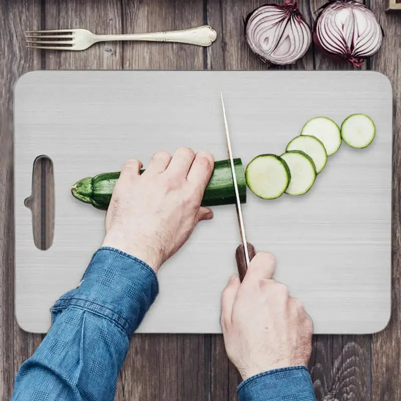 Planche à découper en acier inoxydable, double face épaisse Nesty, mains découpant une courgette en rondelles sur la planche, oignons rouges et fourchette à proximité