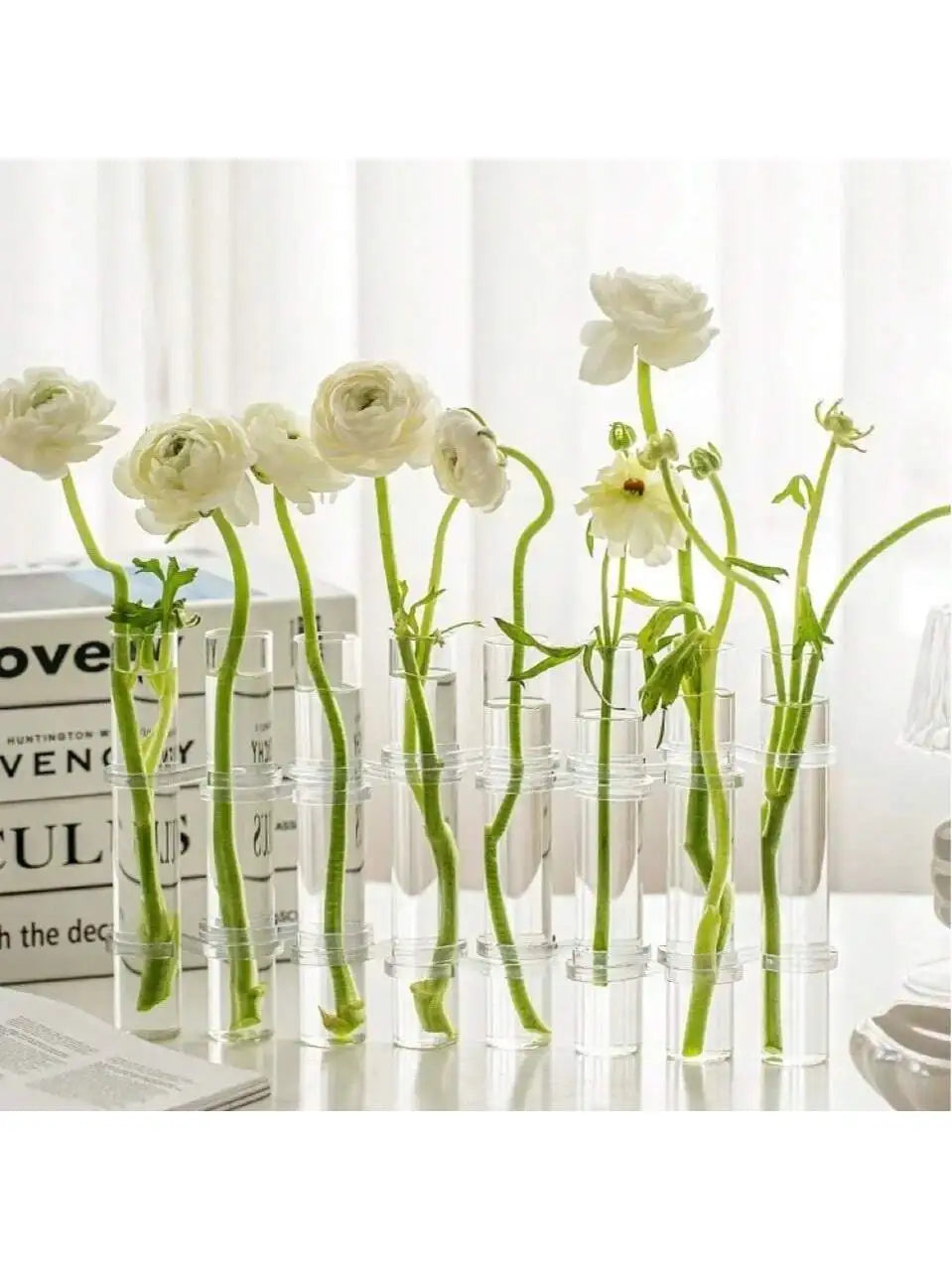 Vase suspendu en verre pour bouturage, lot de tubes transparents Nesty, tubes garnis de renoncules blanches, sur table blanche avec livres