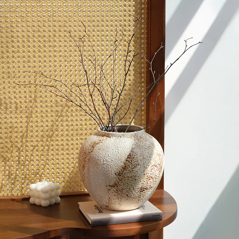 Jarre en céramique artisanale, style wabi-sabi Nesty, format rond à glaçure tachetée, garni de brindilles nues, devant un panneau en cannage