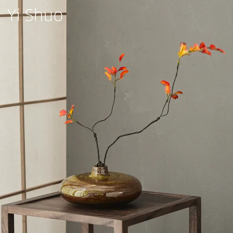 Petit vase en céramique antique Nesty - Forme galet aplati brun-vert, branche fleurie orange, sur table basse en bois