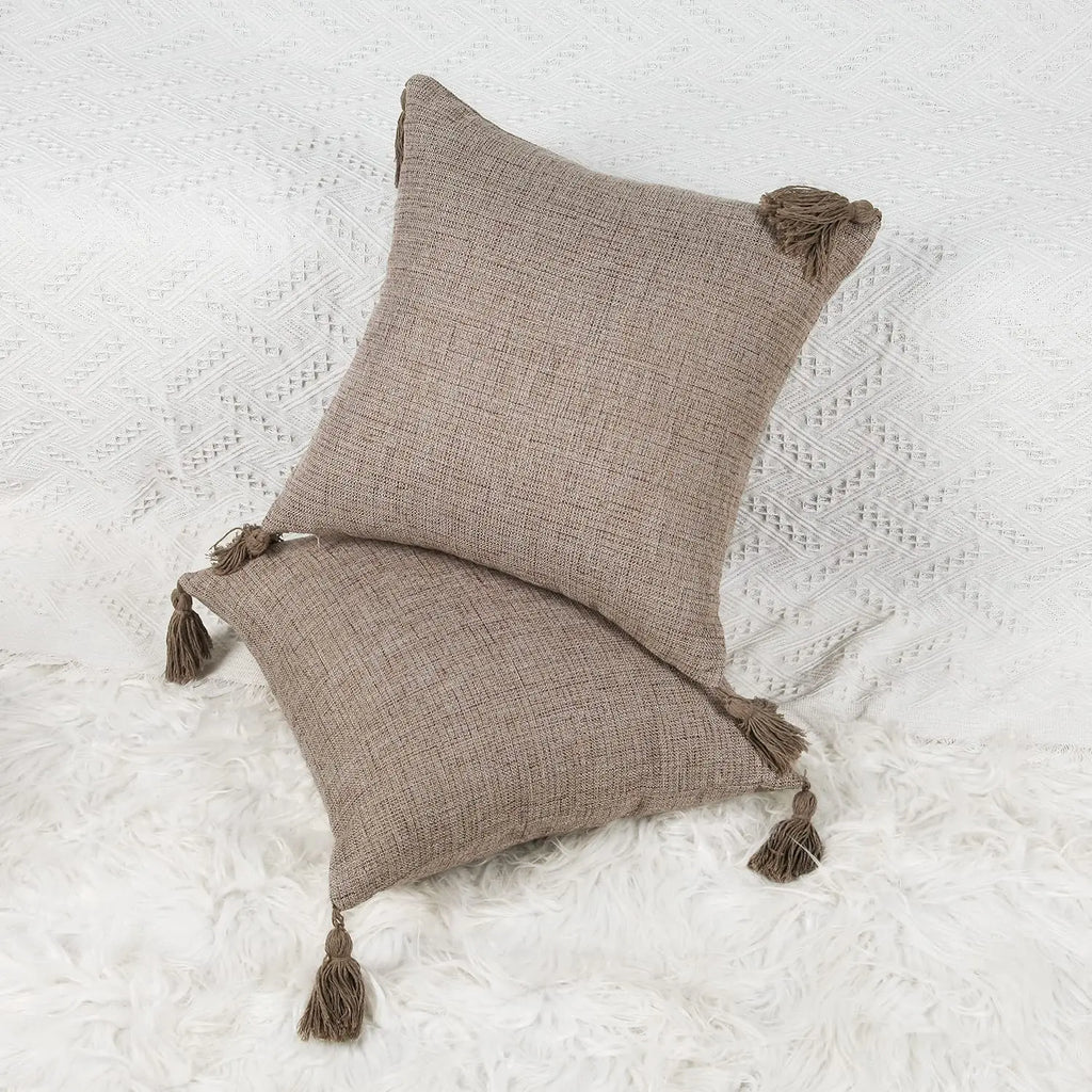 Housse de coussin en lin avec pompons, toutes saisons Nesty, deux housses de coussin marron clair à pompons assortis, empilées sur plaid blanc