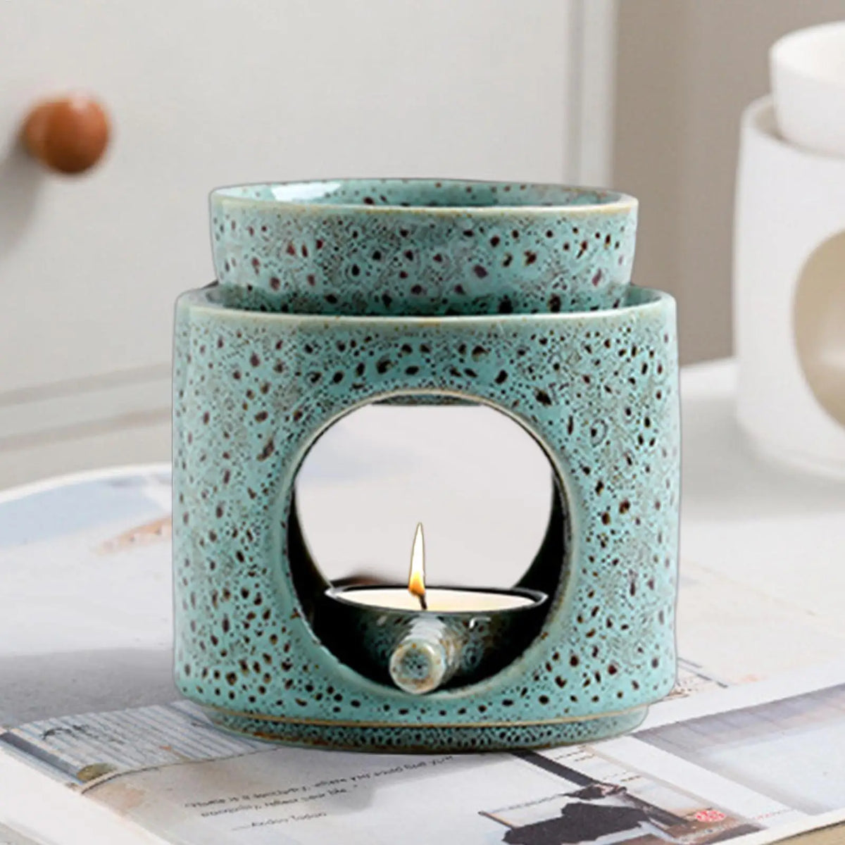 Photophore et chauffe-huile en céramique Nesty - Brûleur turquoise à effet réactif avec coupelle amovible, bougie chauffe-plat allumée à l'intérieur, posé sur un magazine