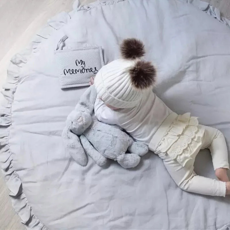 Tapis rond en coton pour bébé — coin jeux - Nesty, variante gris clair à bordure volantée, bébé en bonnet à pompons allongé avec une peluche lapin