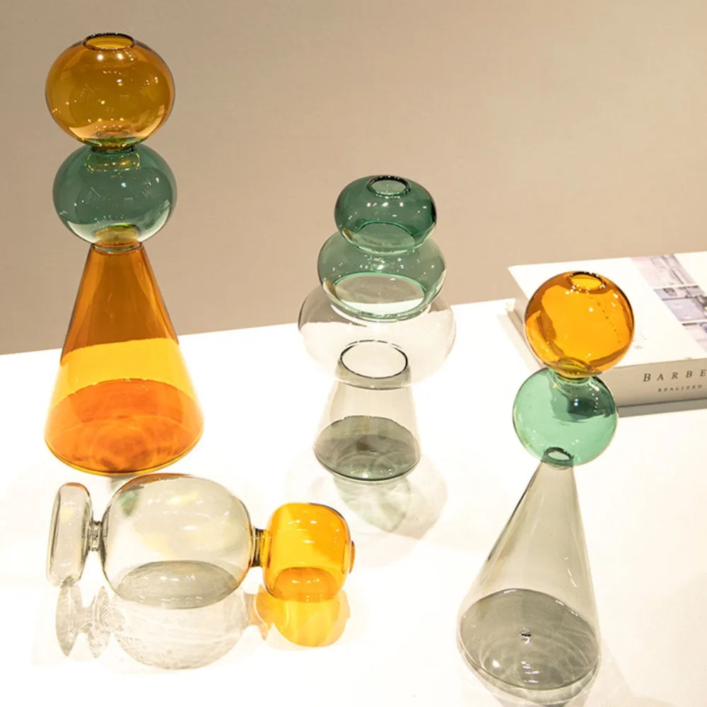 Vase en verre double paroi coloré, culture hydroponique - Nesty - Cinq modèles aux coloris ambré, vert et gris disposés en groupe sur une table blanche près d'un livre