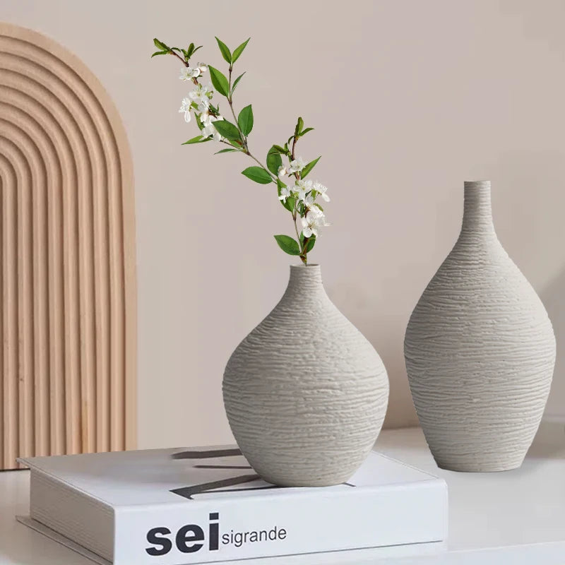 Vase en céramique à col étroit, style nordique sobre Nesty, deux vases en céramique texturée forme bouteille gris clair, l'un avec une branche fleurie, posés sur un livre "sei sigrande" devant une arche en bois