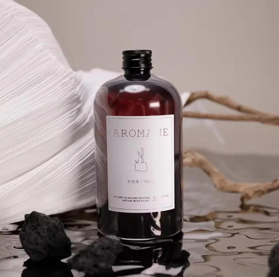 Diffuseur de parfum d'ambiance Nesty - Flacon ambré AROMA'ME posé sur galets noirs et branche de bois flotté